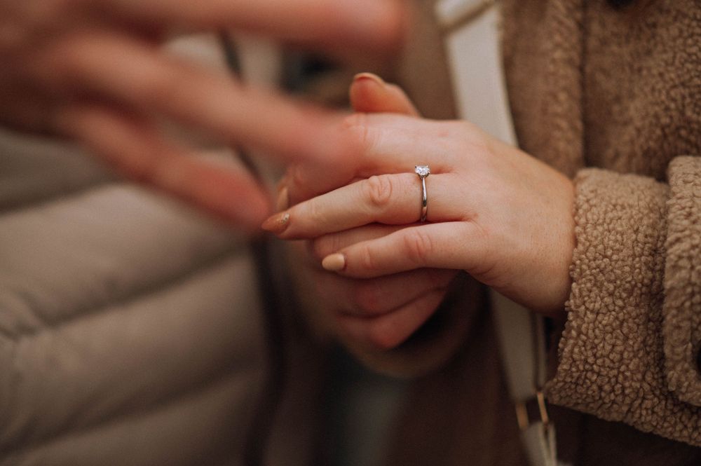 Close-up of a woman's hand wearing a solitaire diamond engagement ring, held gently by another person.
