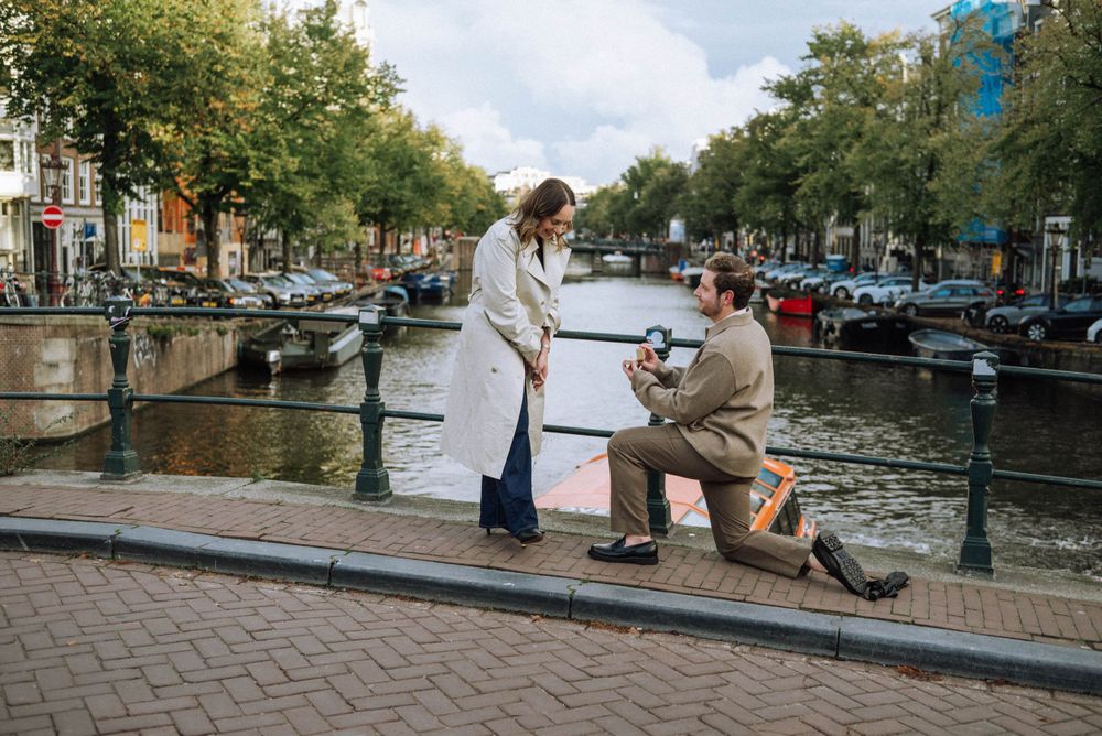 A man on one knee, holding a ring in a heartfelt secret proposal moment on one of Amsterdam’s historic bridges, with a canal in the backgrou