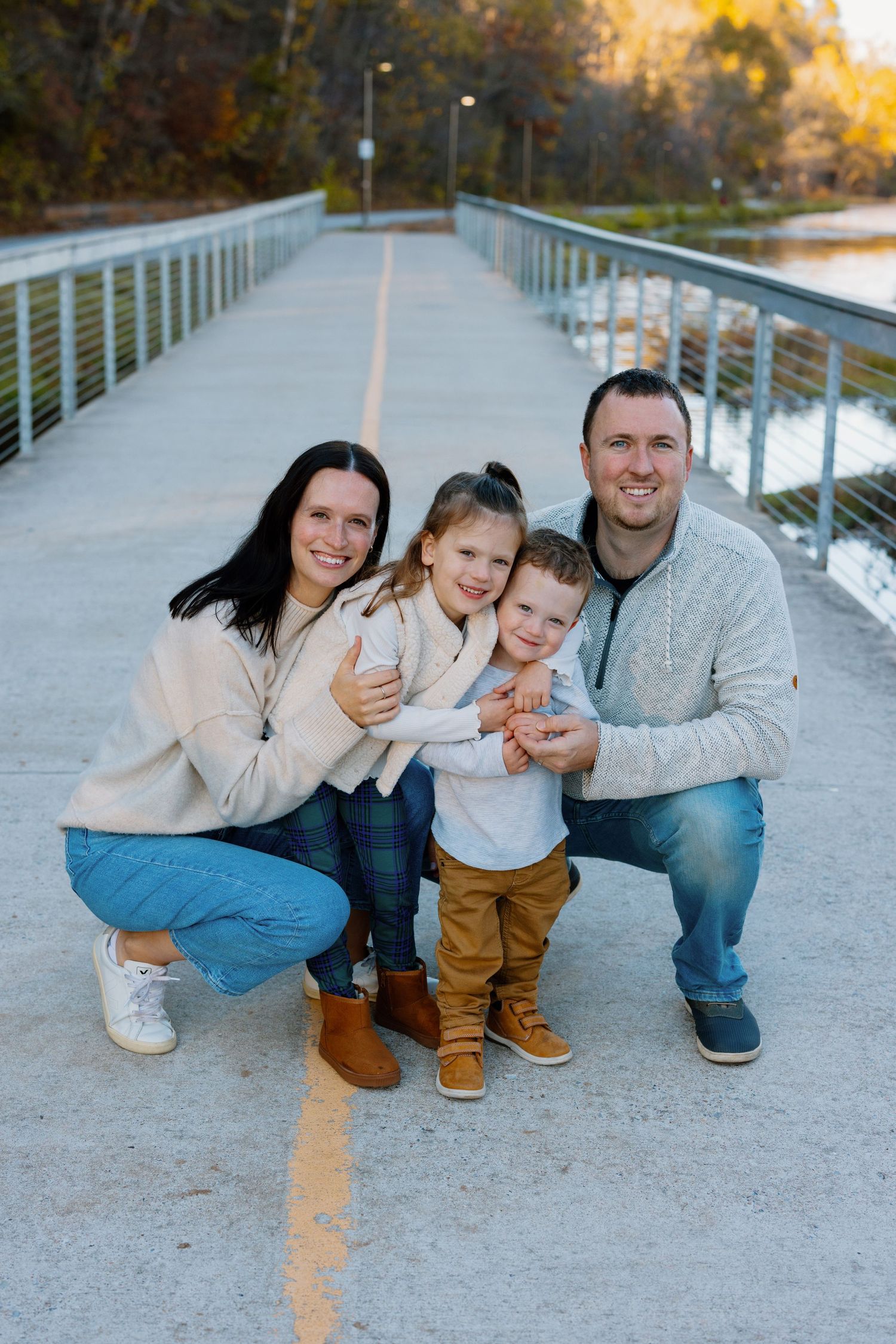 Family poses together on a bridge walkway during autumn with golden trees in the background.
