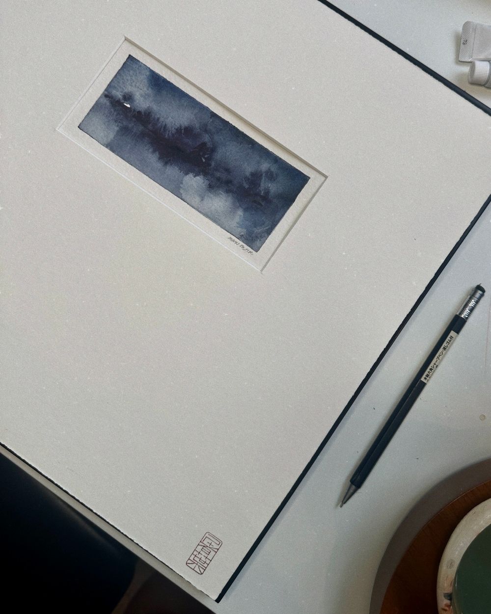 Small watercolor painting of a dark misty landscape mounted on white paper with a pen sitting beside it on the desk.