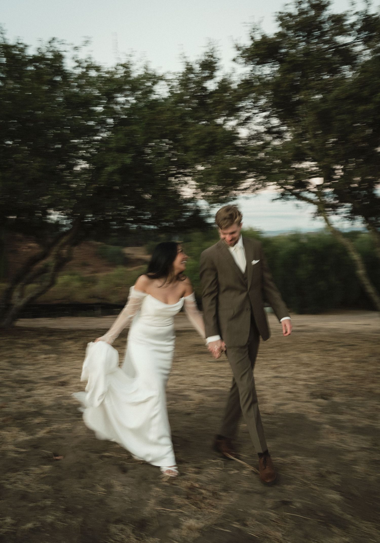 A couple in wedding attire walks hand in hand through a scenic outdoor setting with trees in the background.