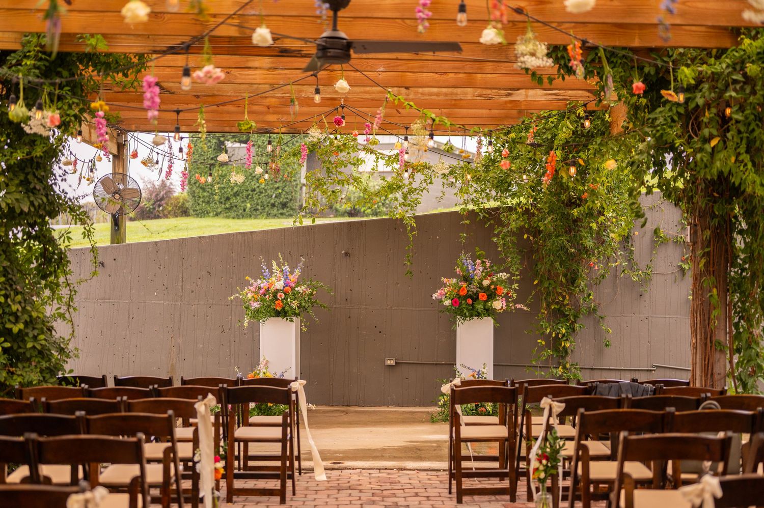Empty rows of wooden chairs set up for outdoor wedding ceremony under wooden canopy with string lights and hanging floral decor.