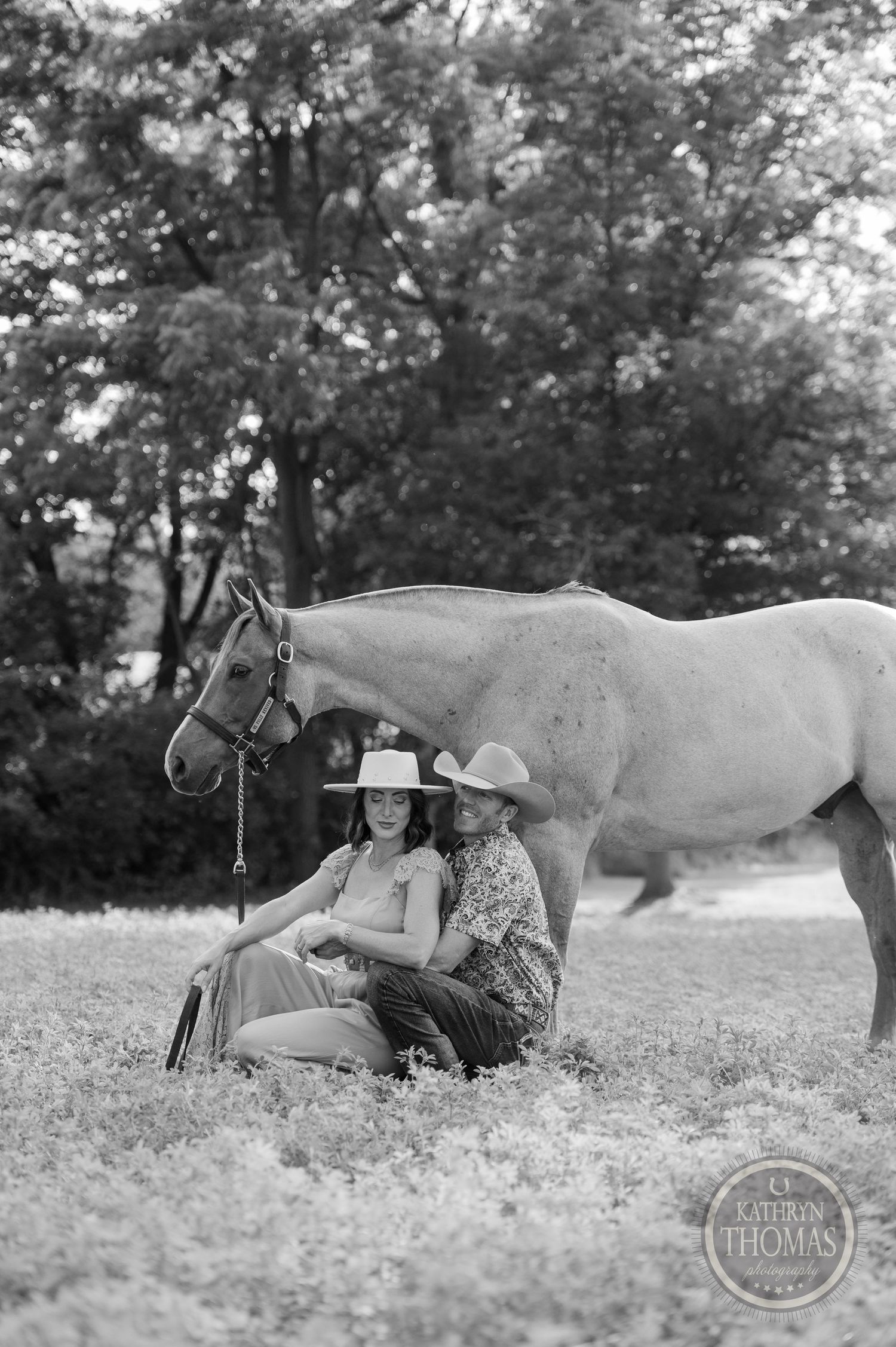 A black and white photograph of a white horse standing with people relaxing in a grassy field.