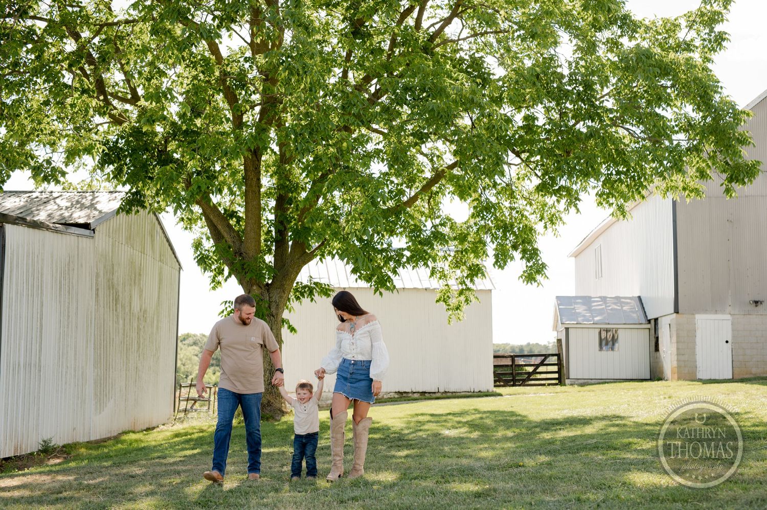 Family walking hand in hand under large tree with white barn in background.