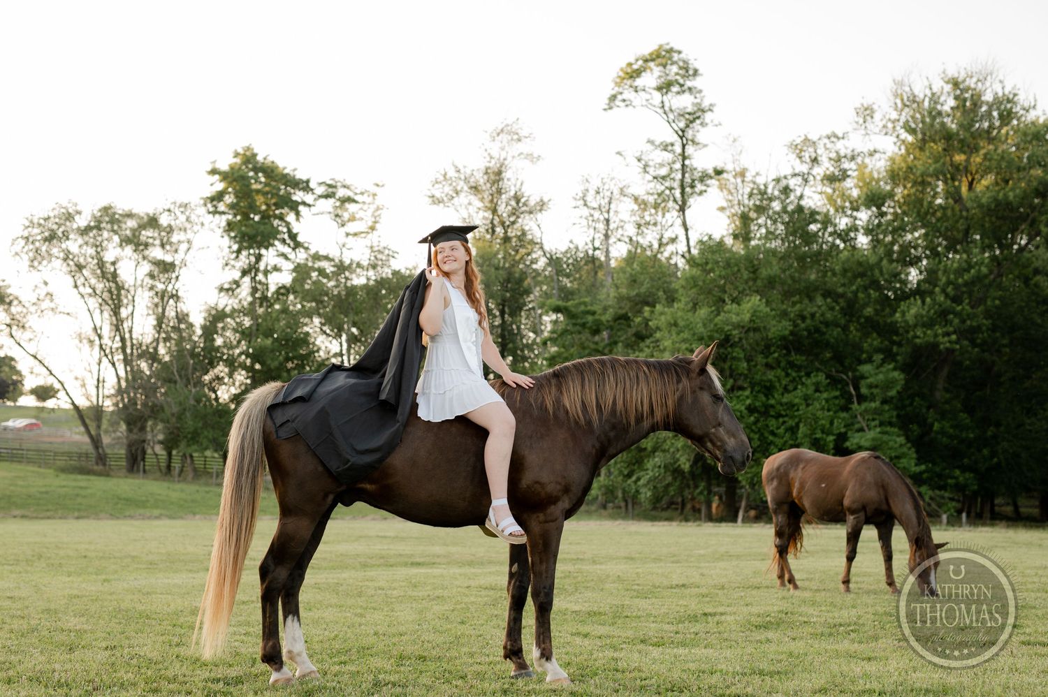 A graduate in cap and gown poses on horseback while another horse grazes in the background.
