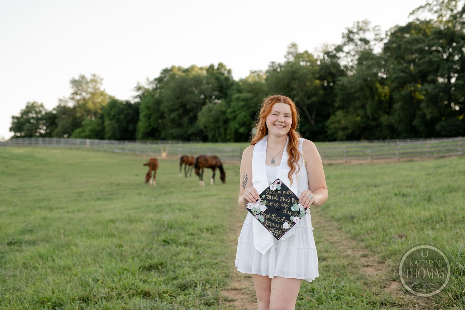 Graduate in white dress holds bouquet in field with horses grazing in background.