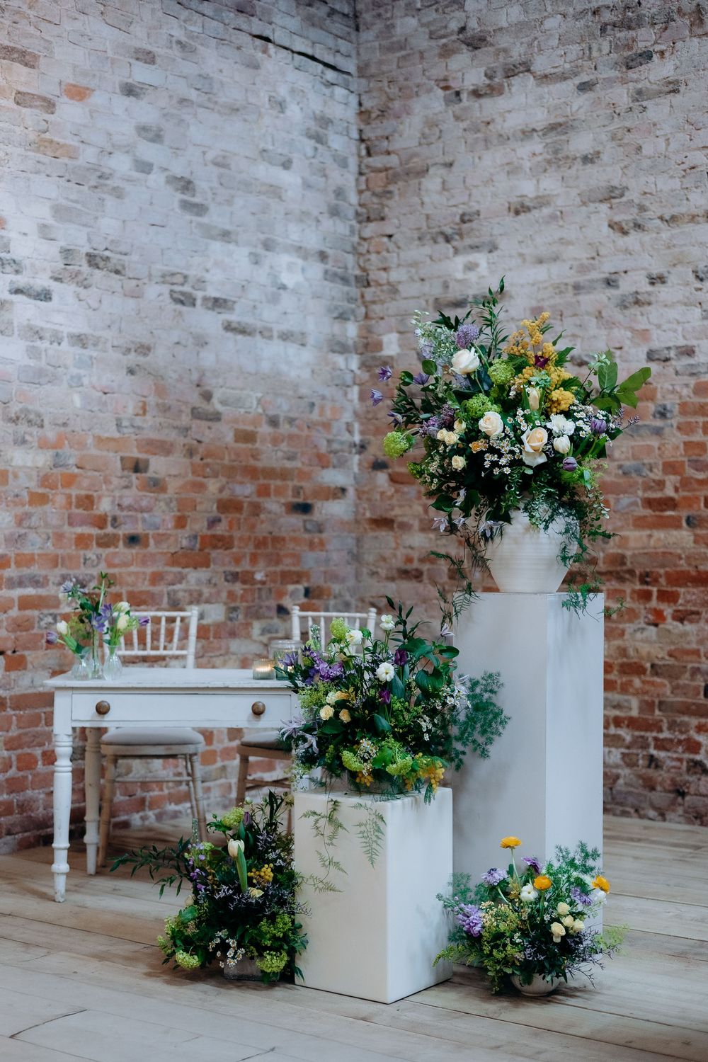 White floral arrangements with greenery displayed against rustic brick walls in an indoor venue setting.