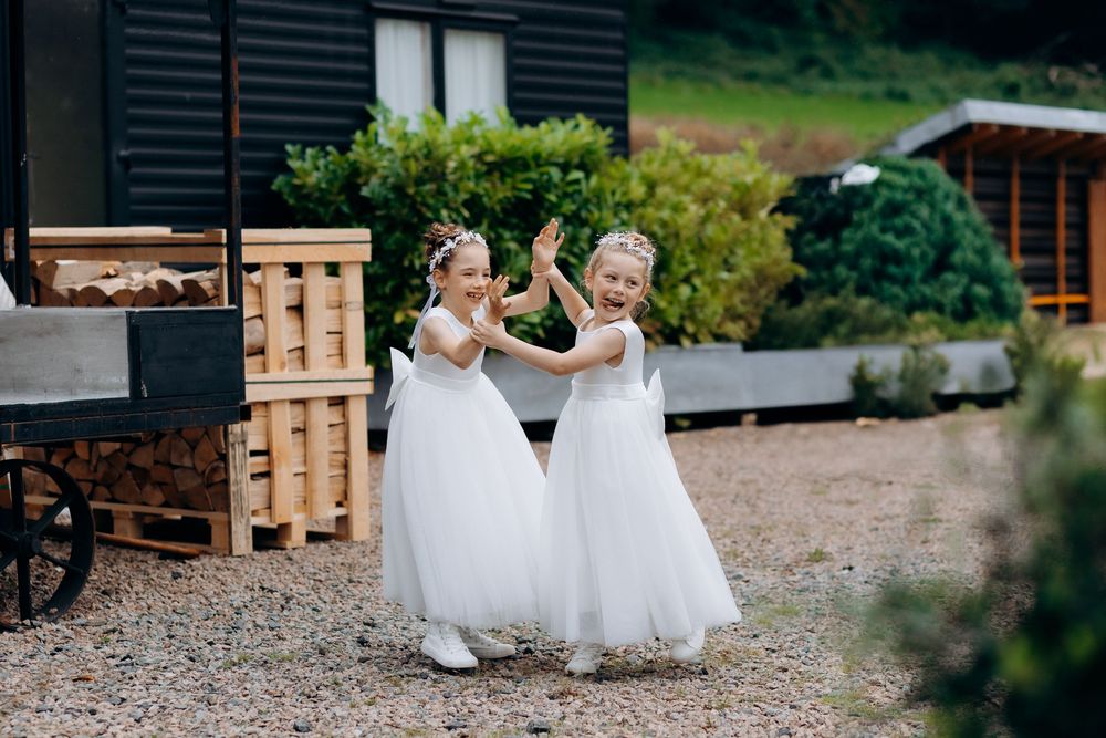Two people in white dresses sit together on rustic wooden benches in an outdoor garden setting.