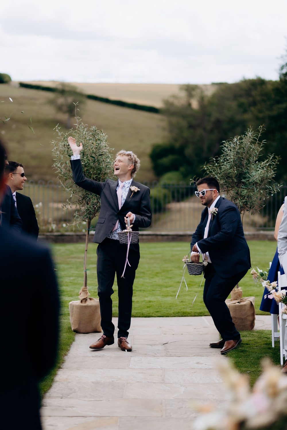 A vibrant wedding celebration with guests in suits jumping and dancing on an outdoor patio with rolling hills in the background.