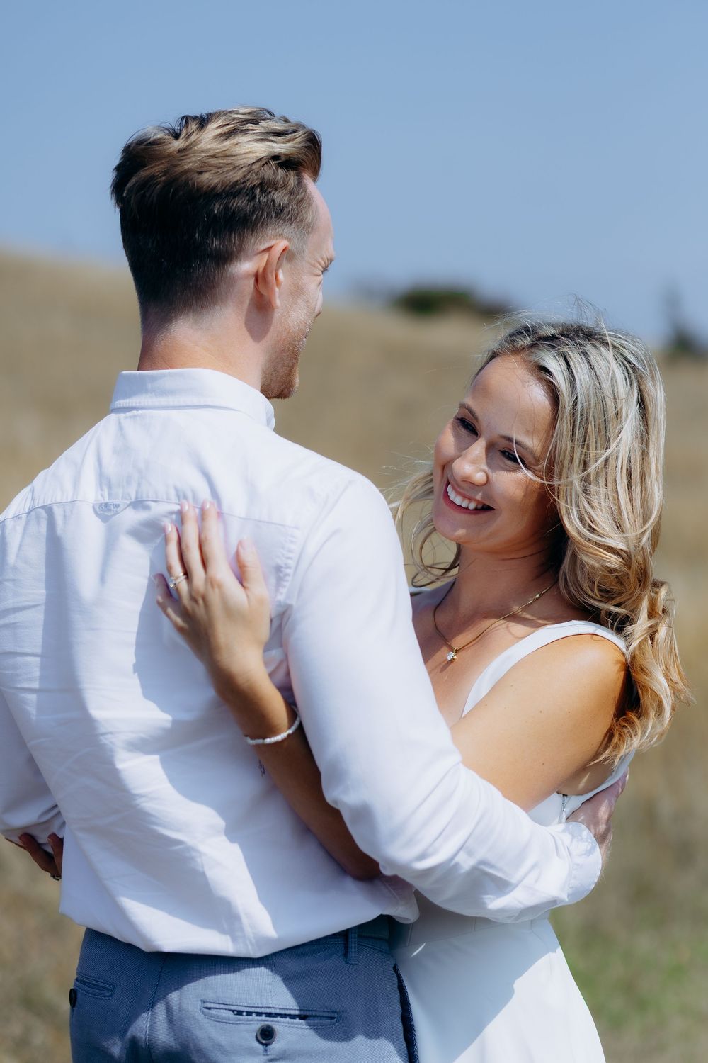 A couple in wedding attire embrace and dance together in a sunny outdoor field.