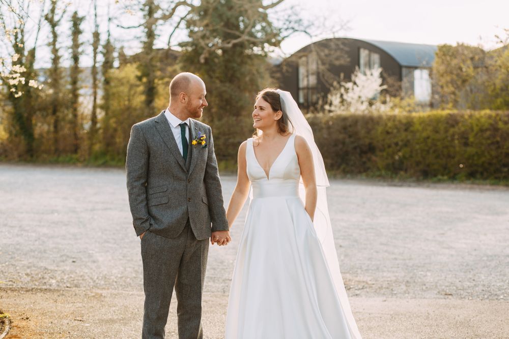 Bride and groom in elegant attire share a moment on gravel path near rustic building.
