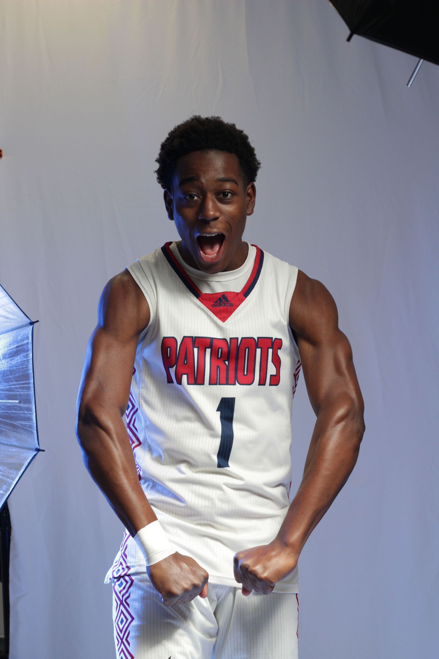 Basketball player in white Patriots jersey number 1 poses energetically against a studio backdrop with lighting umbrellas visible.
