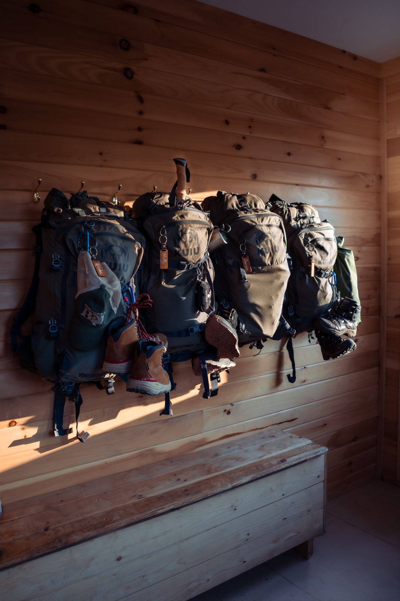 Multiple rugged backpacks and bags hanging on wooden wall hooks in a cabin or lodge with dramatic lighting.