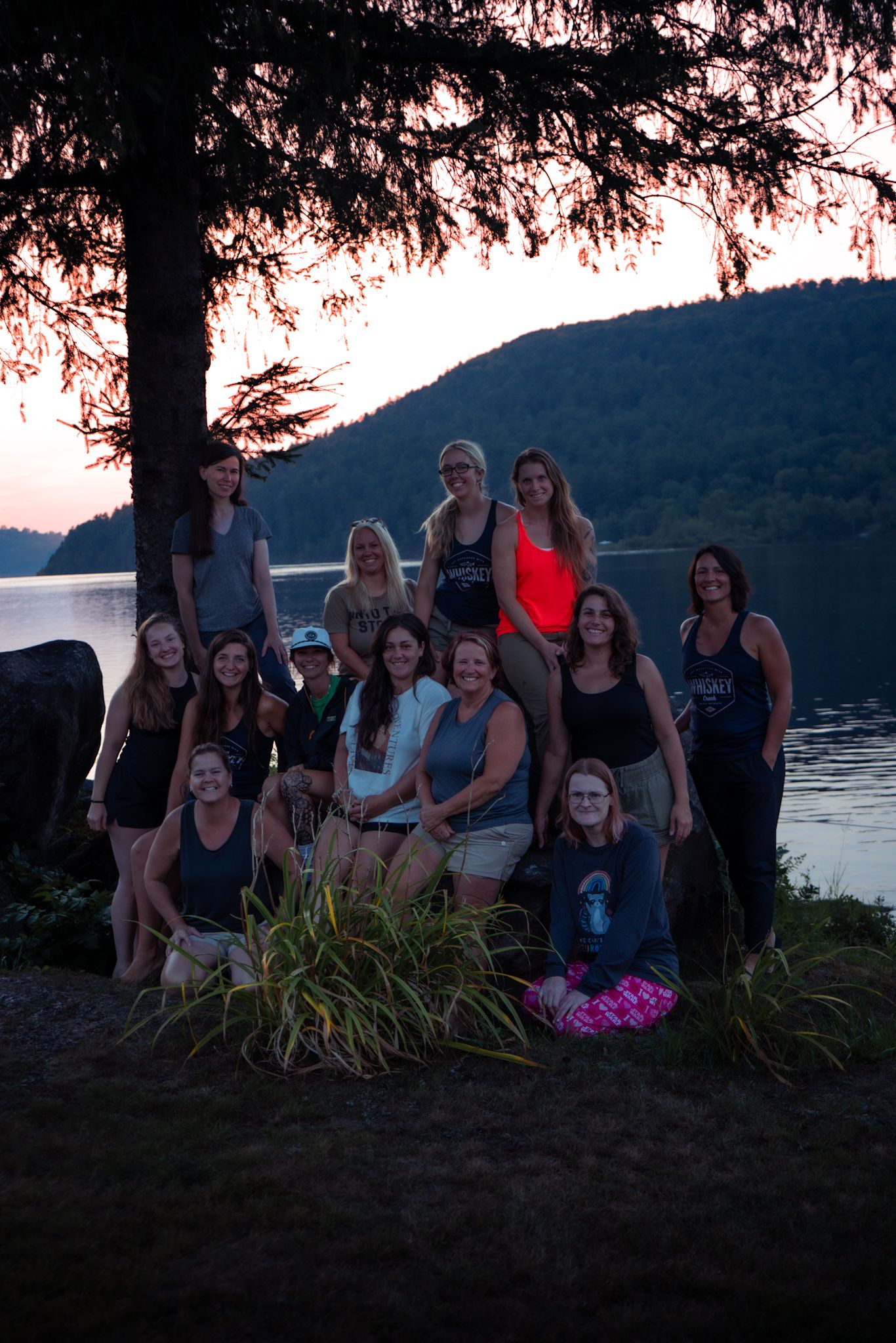 Group gathered at sunset by a lakeside with mountains in background under a tree's silhouette.