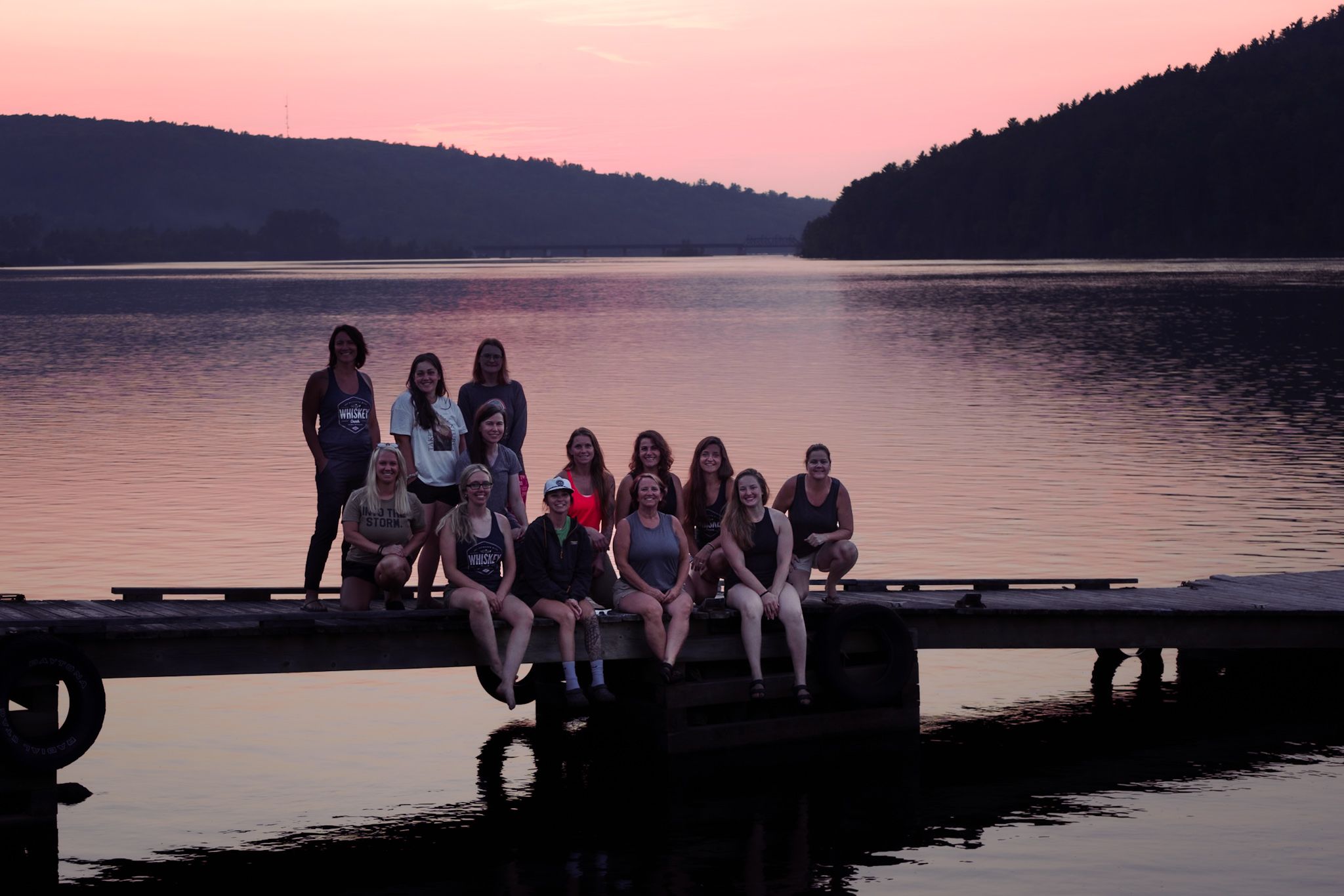 A group sits together on a wooden dock at sunset, silhouetted against a peaceful lake with forested hills in the background.