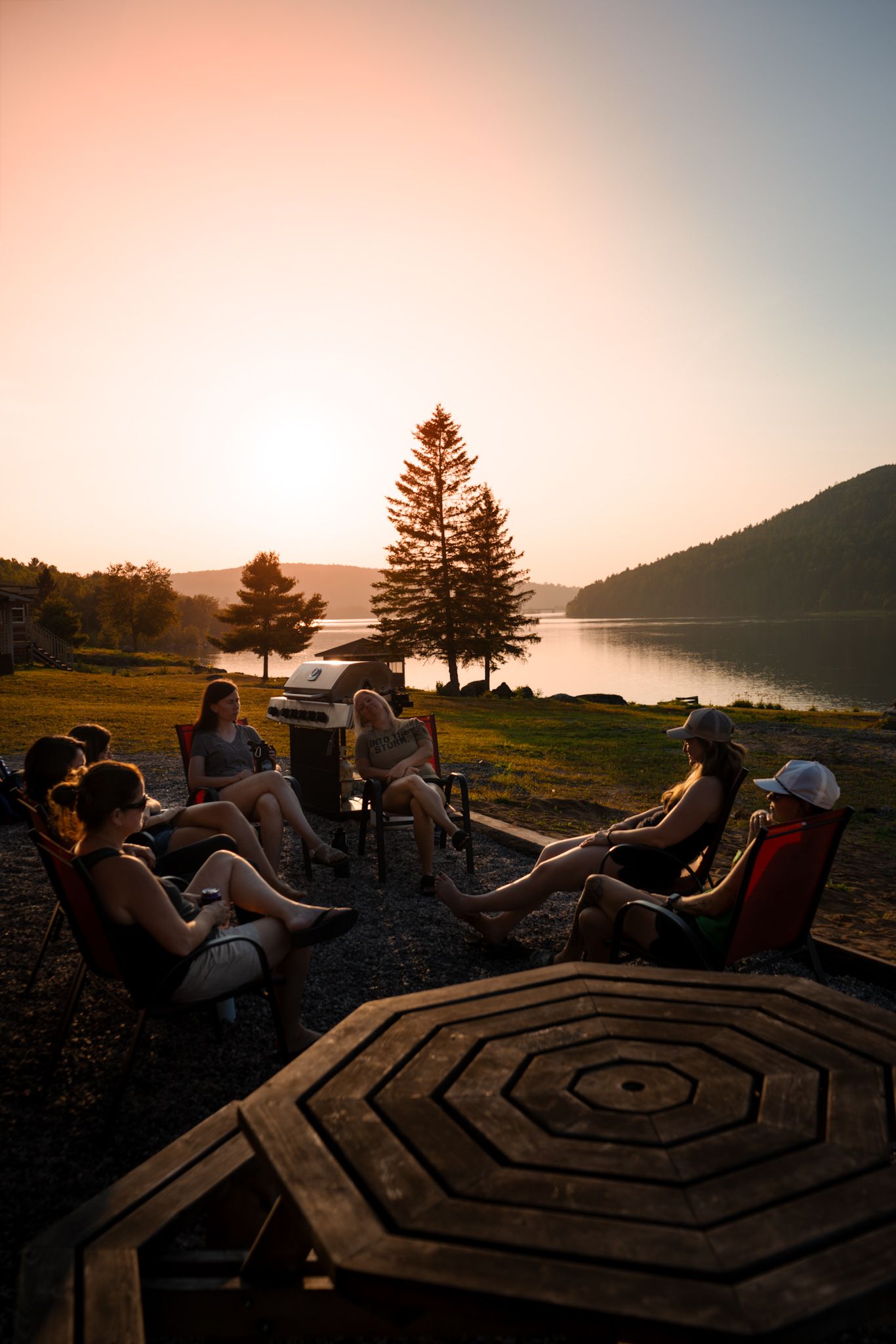A group gathers around a campfire table at sunset by a serene lake with pine trees silhouetted against the pink sky.
