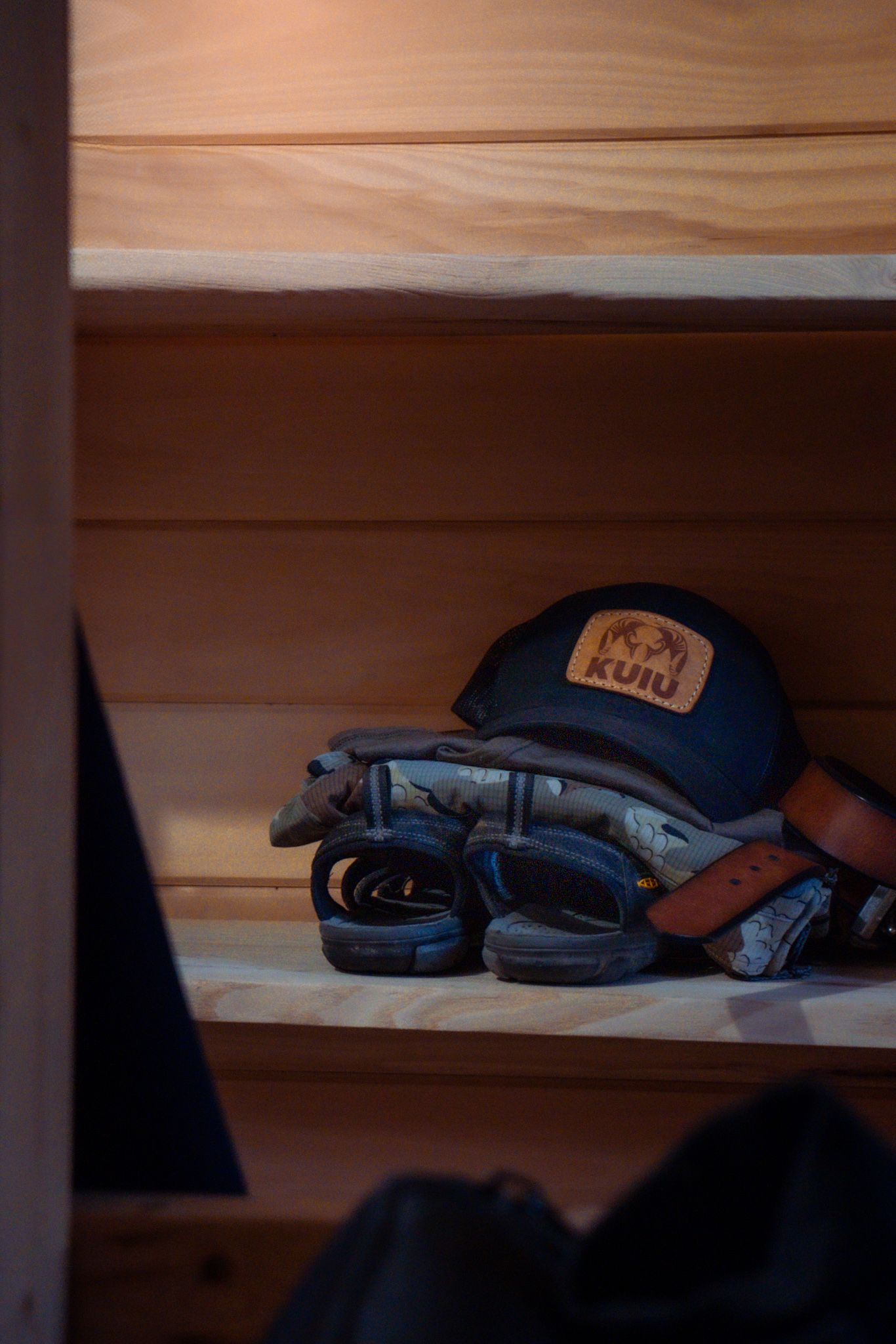 A blue baseball cap and hiking sandals sit on a wooden shelf in dim lighting.
