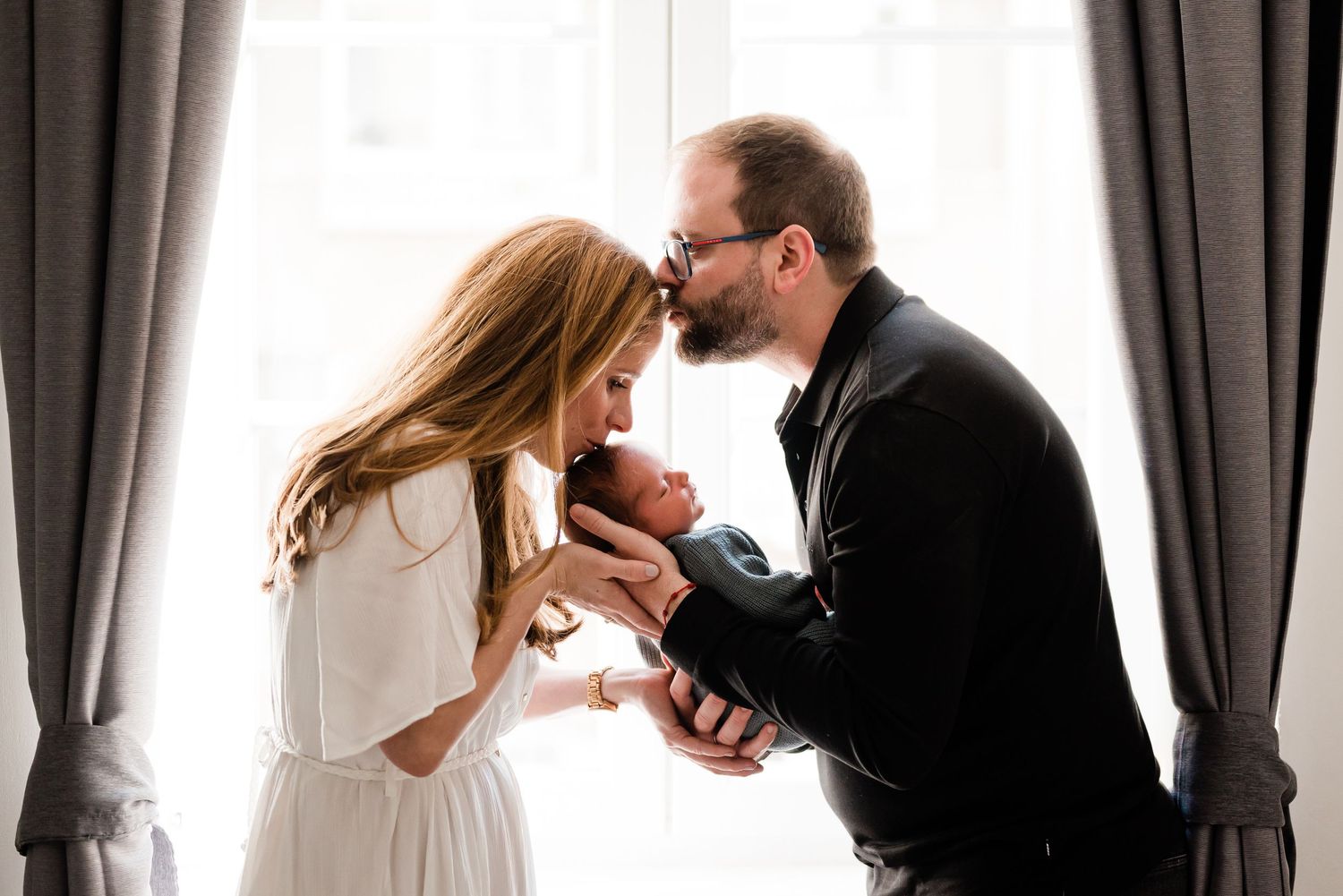 A mother and father loving kiss their newborn, bathed in soft natural light by a window framed by grey curtains at home in Nyon, Switzerland