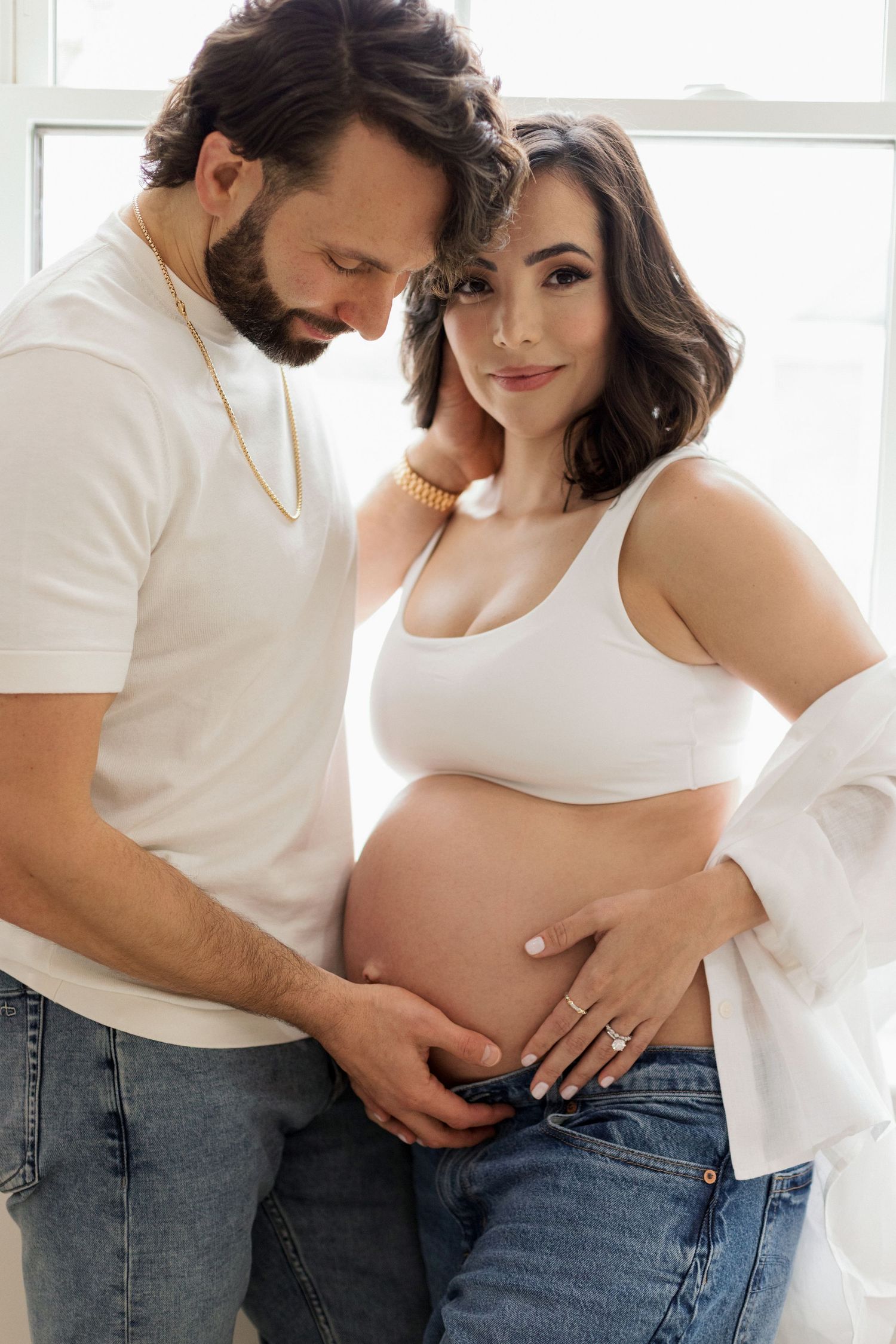 A serene maternity moment by a bright window in winter, wearing a cozy white sweater dress against natural light.