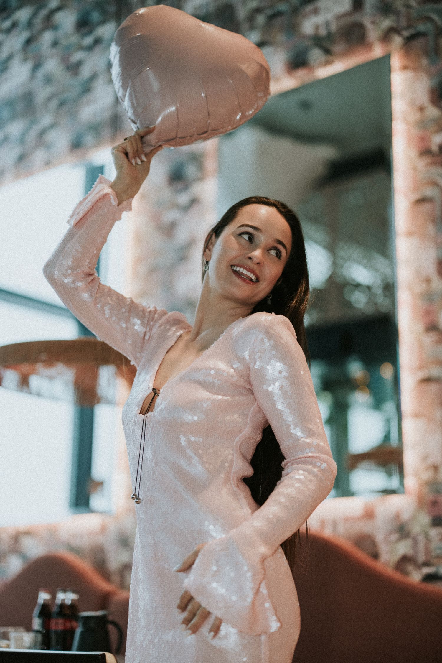 A woman in a sequined pink dress holds a heart-shaped balloon overhead while posing in an elegant venue.