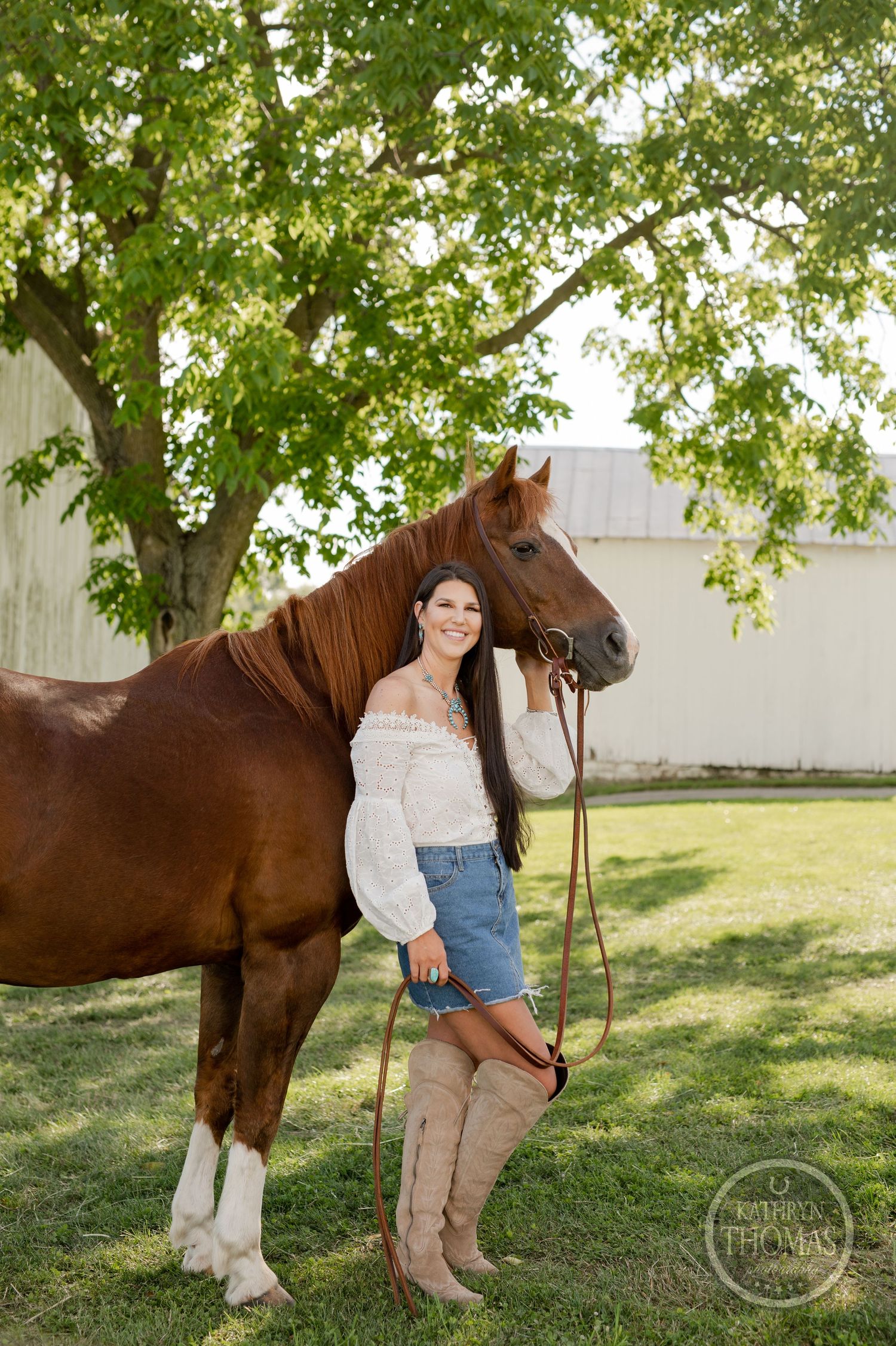 A chestnut horse with white legs poses next to a rustic white barn under a shady tree.