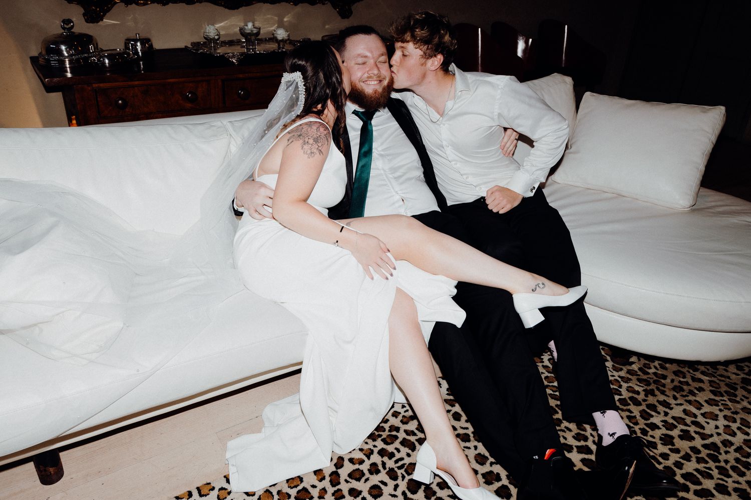The bride and a groomsman kissed the groom on an elegant couch, captured by Apertured Media Auckland Wedding Photographer.