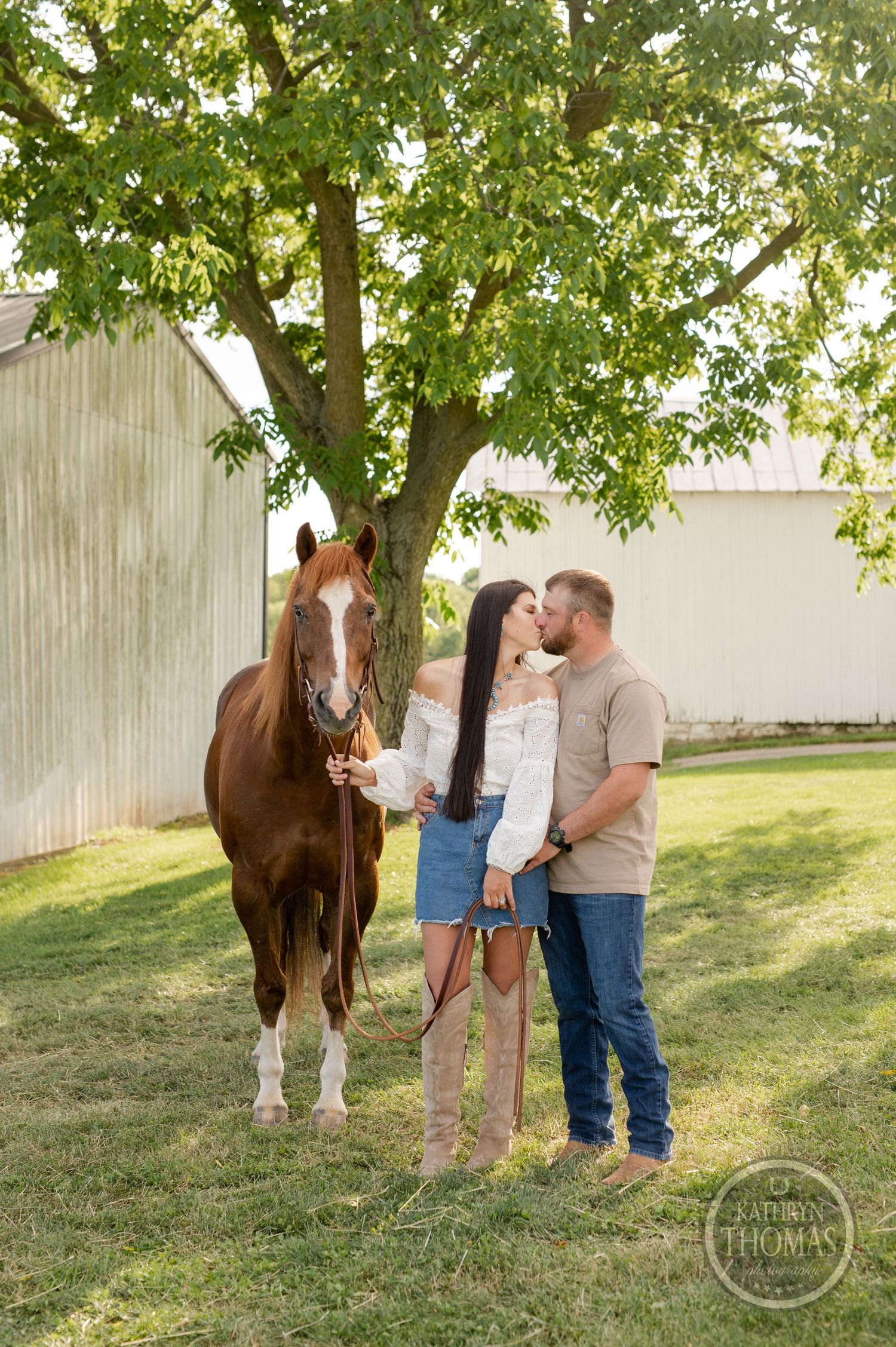 Couple embraces near brown horse under large tree by white barn wall.