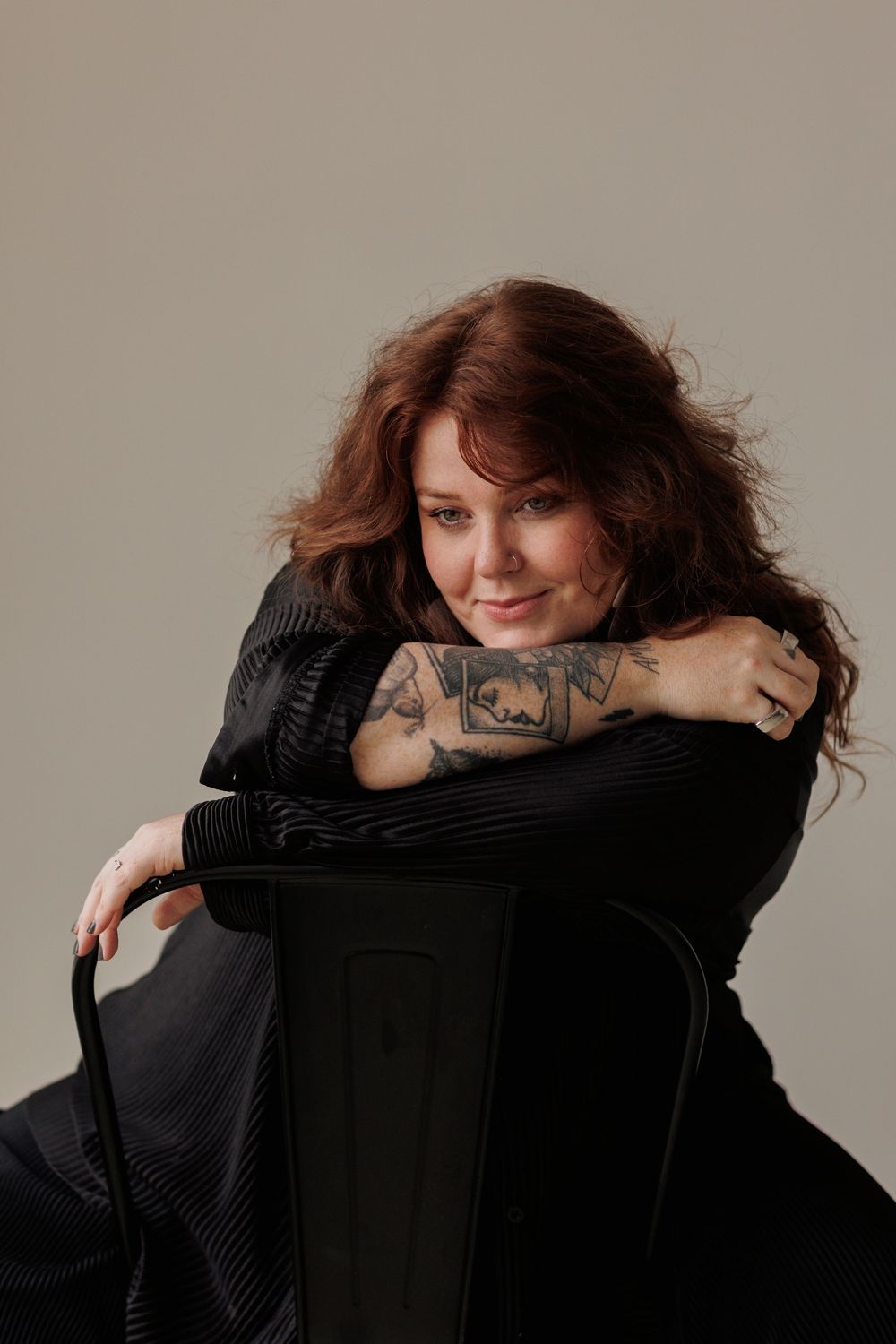 A person with long wavy auburn hair and tattoos poses on a black chair against a neutral backdrop.