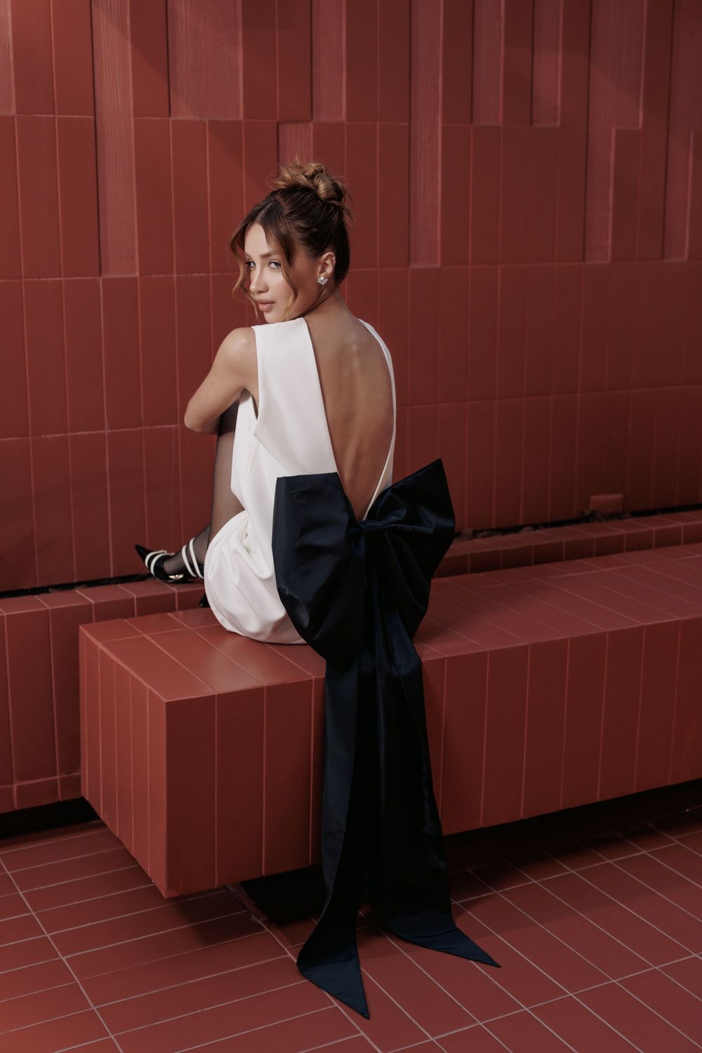 Elegant model in white backless dress with dramatic black bow detail poses on red tiled steps.
