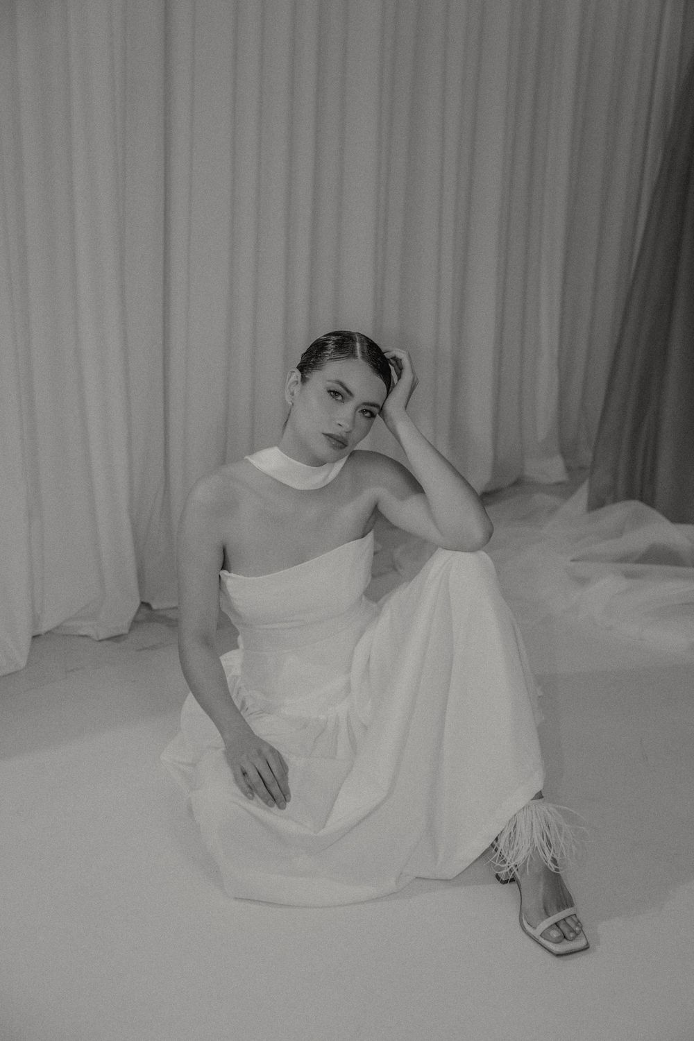 Black and white portrait of someone in an elegant white one-shoulder dress and heels sitting against a light backdrop.