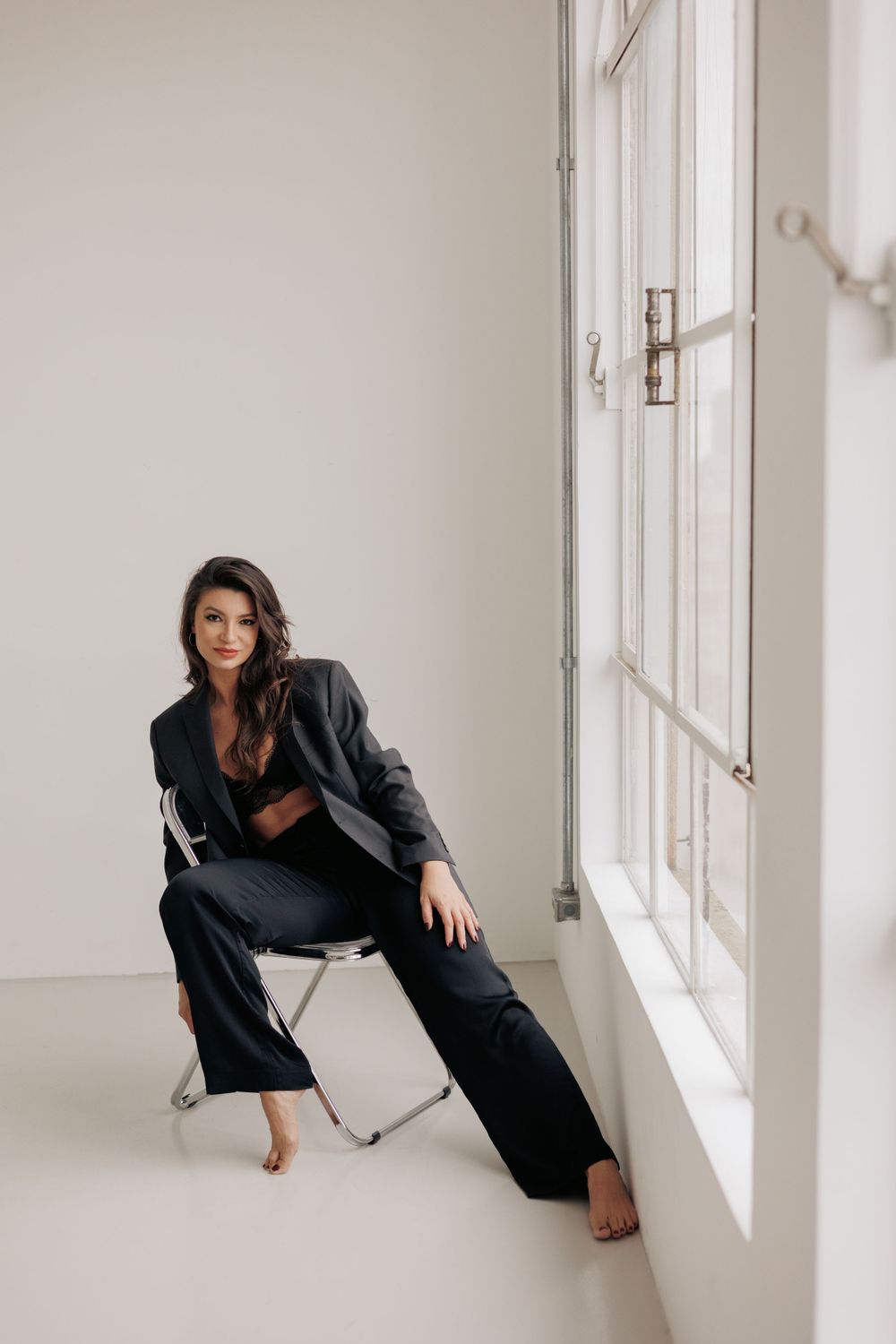 A person in dark loungewear sits casually on a folding chair by large windows.