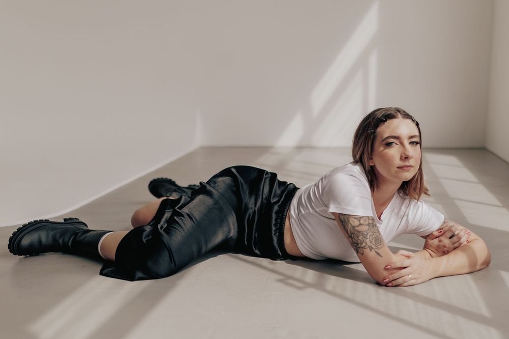 Person in white t-shirt and dark jeans lying on floor near white wall with natural lighting and shadows.