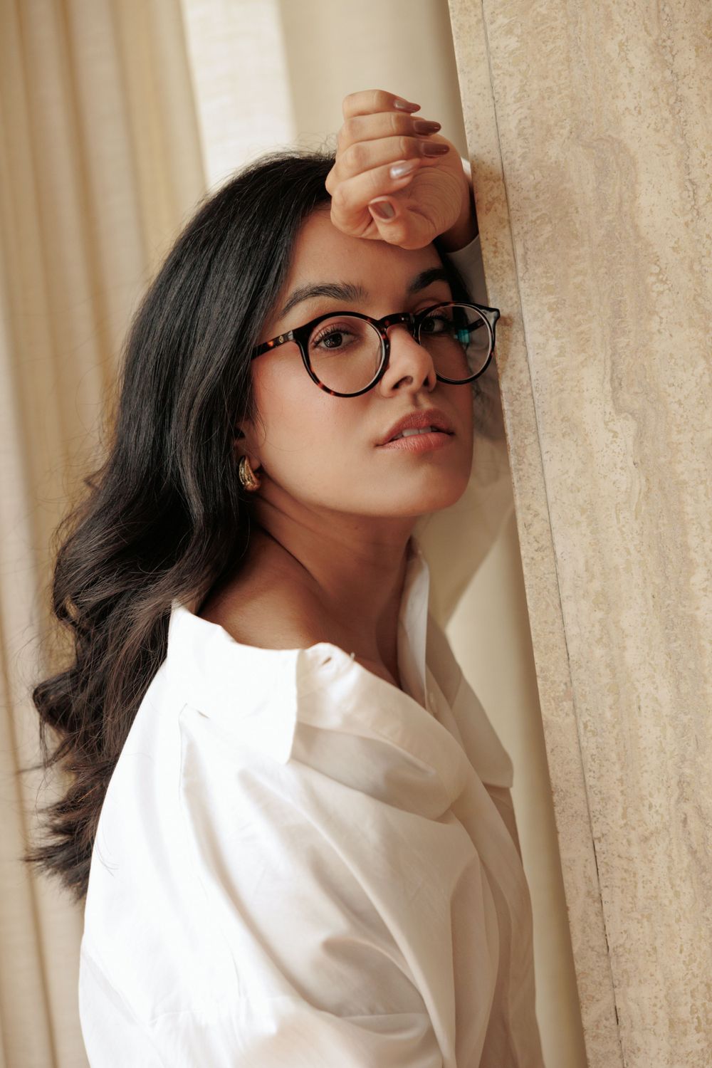 Portrait of woman in glasses and white shirt leaning against curtained wall.