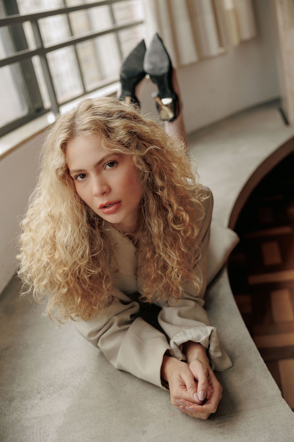 Fashion portrait of a woman with blonde curls lying on a windowsill in natural light.