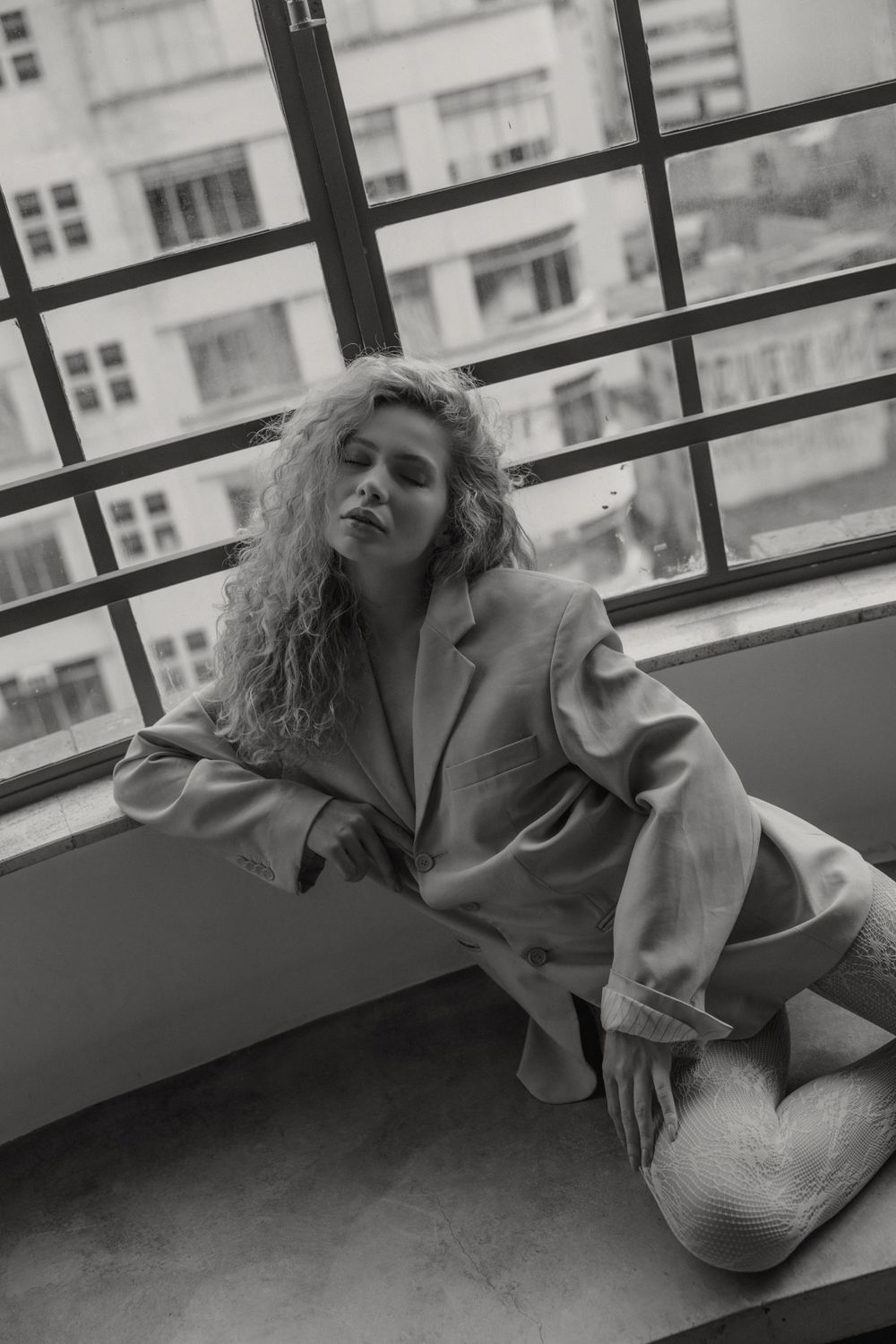 Black and white photo of person in oversized blazer reclining near urban window view.