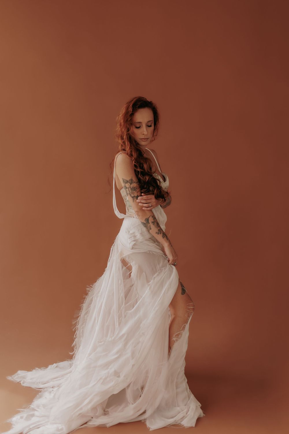 A graceful figure in a flowing white wedding dress poses elegantly against a warm brown backdrop in a studio setting.
