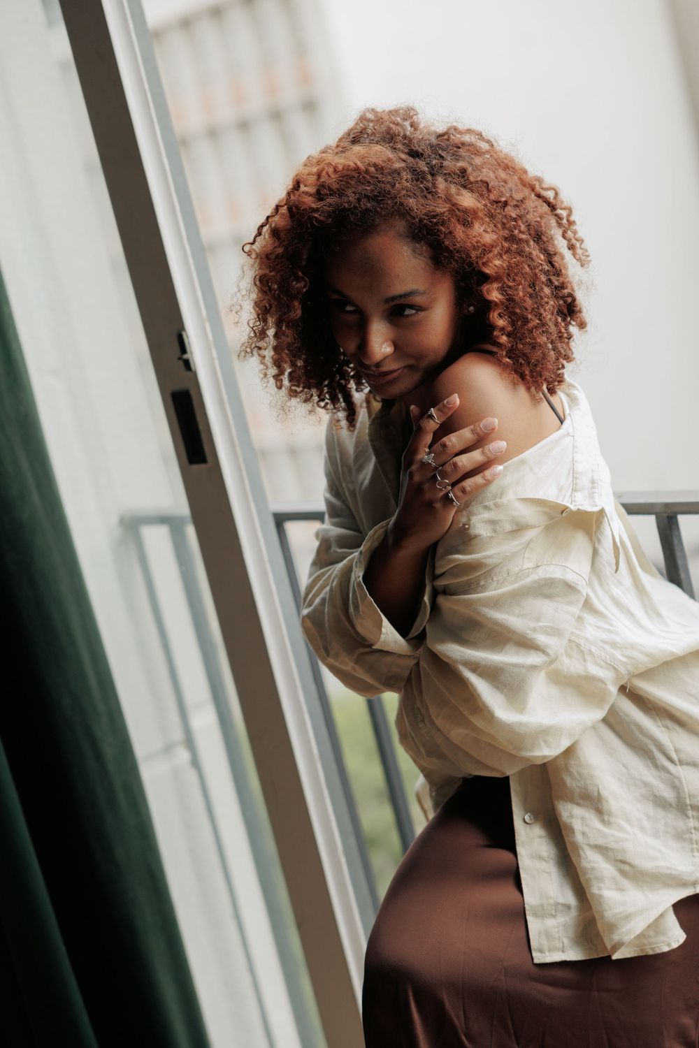 Woman with natural curly hair poses playfully near window in casual cream shirt.
