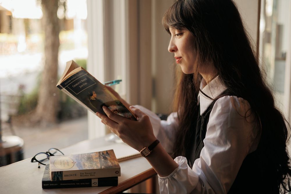 Two people sit side by side on a couch near a bright window, both absorbed in reading books.