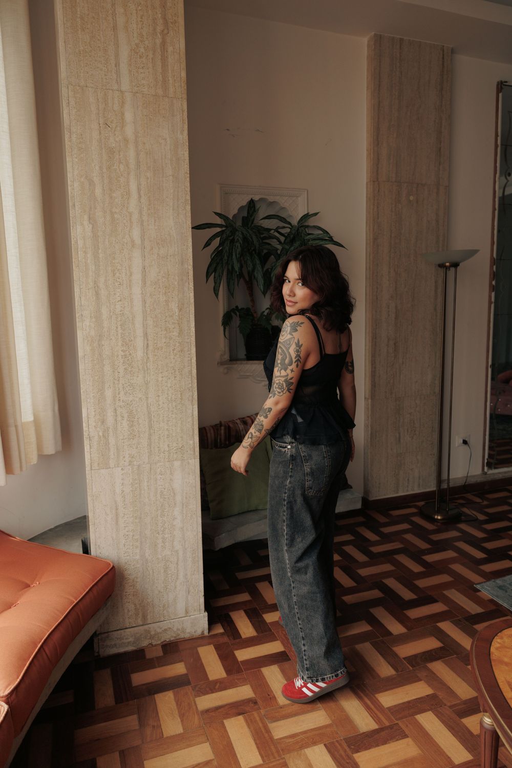 Woman with tattoos poses in black top and jeans against chevron wood flooring.