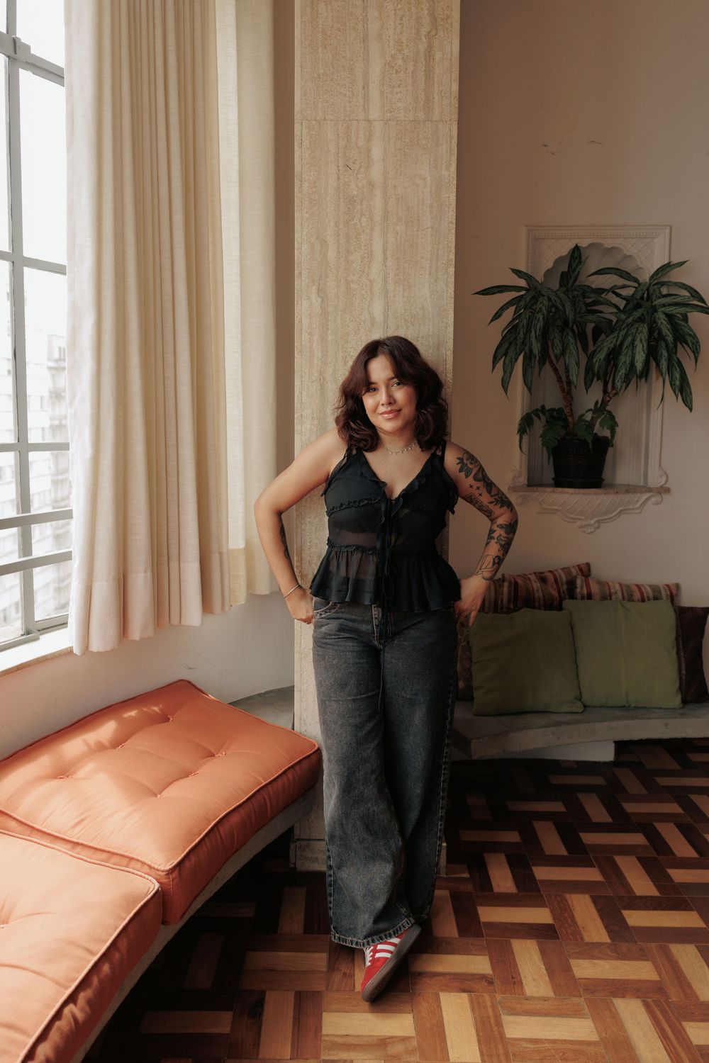 Woman in black top and wide-leg jeans stands confidently by window in modern apartment.