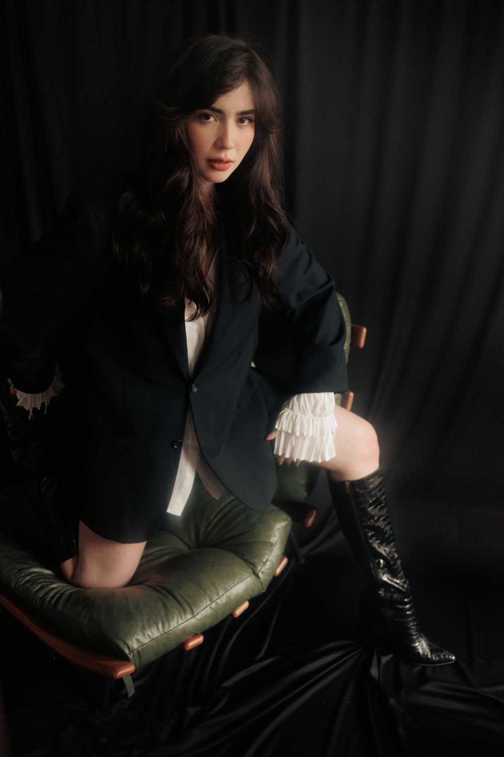 Dark moody fashion portrait with model in black blazer and boots against dark backdrop.