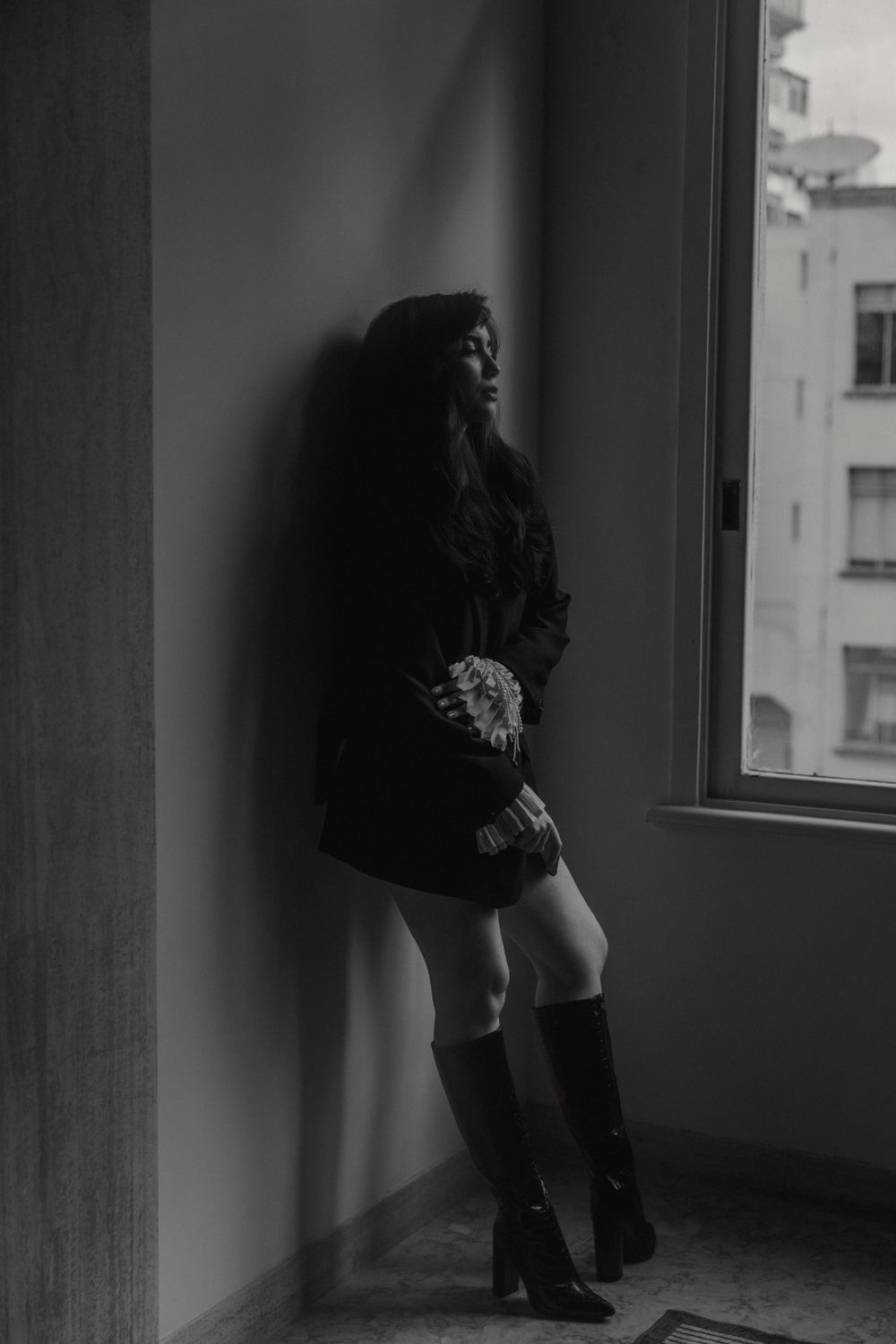 Monochrome portrait of figure in tall boots standing by window in dramatic lighting.