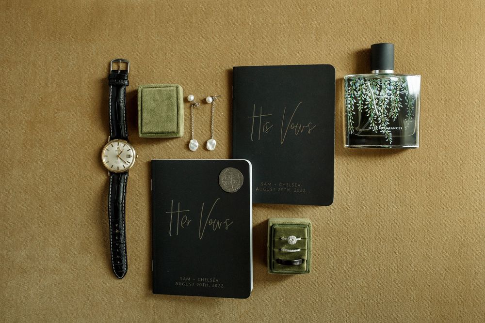 Flat lay of wedding details including watch, jewelry, and black vow books on brown background.