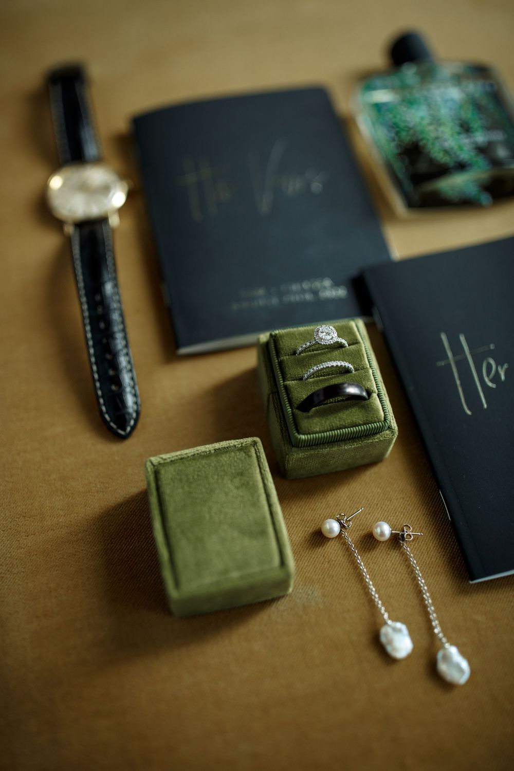 Wedding accessories including rings, watch, earrings and black notebooks arranged on brown surface.