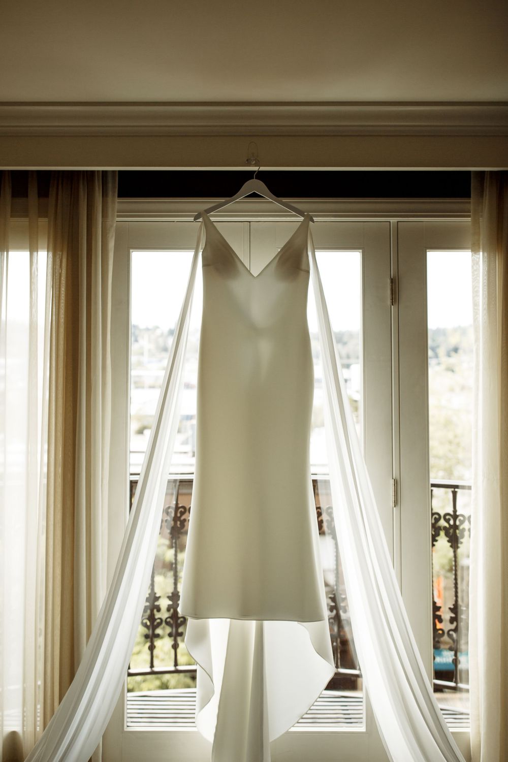 A sleek white wedding dress hangs elegantly in front of French doors with flowing curtains.