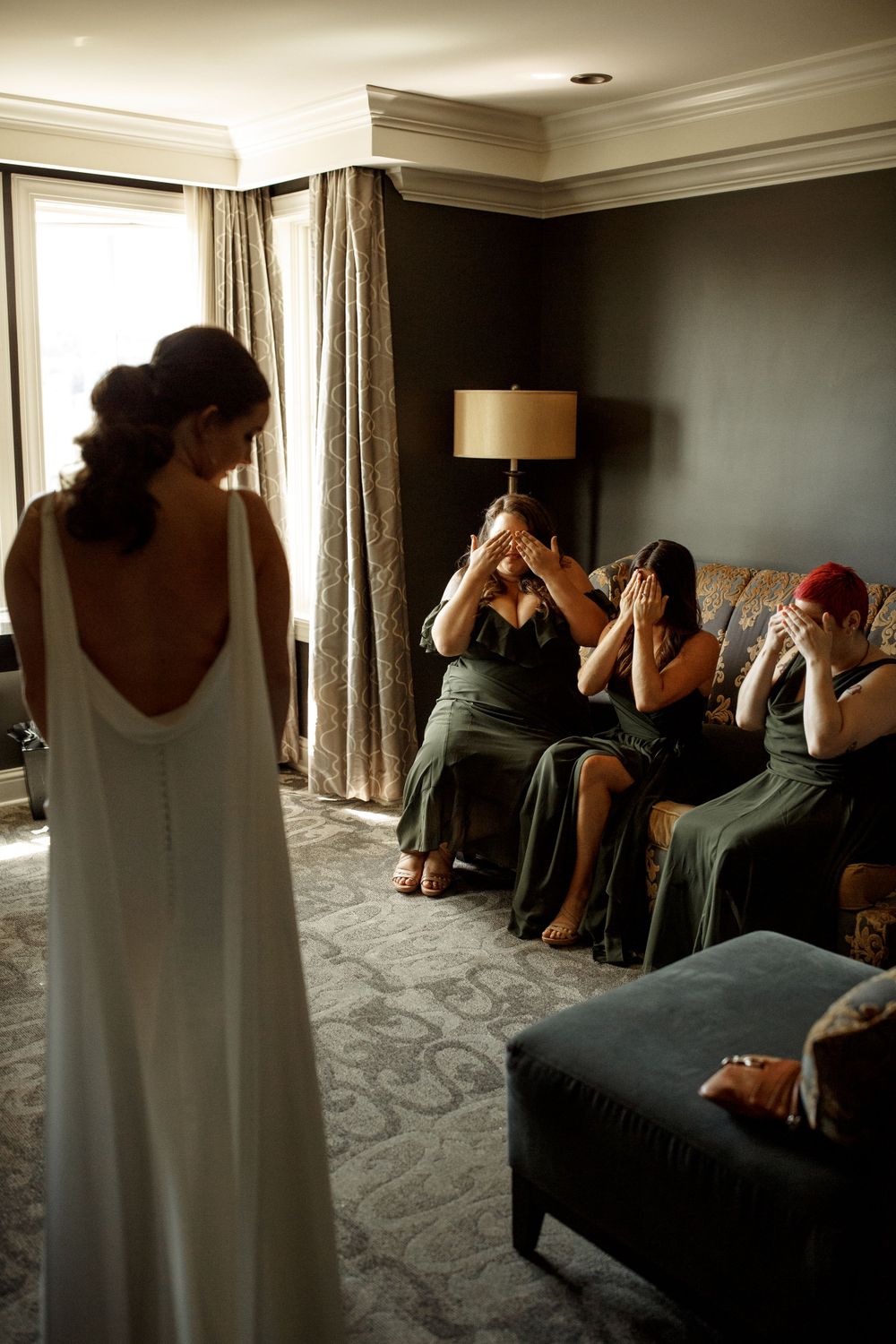 A wedding dress with dramatic low back is displayed as others react emotionally on a couch.