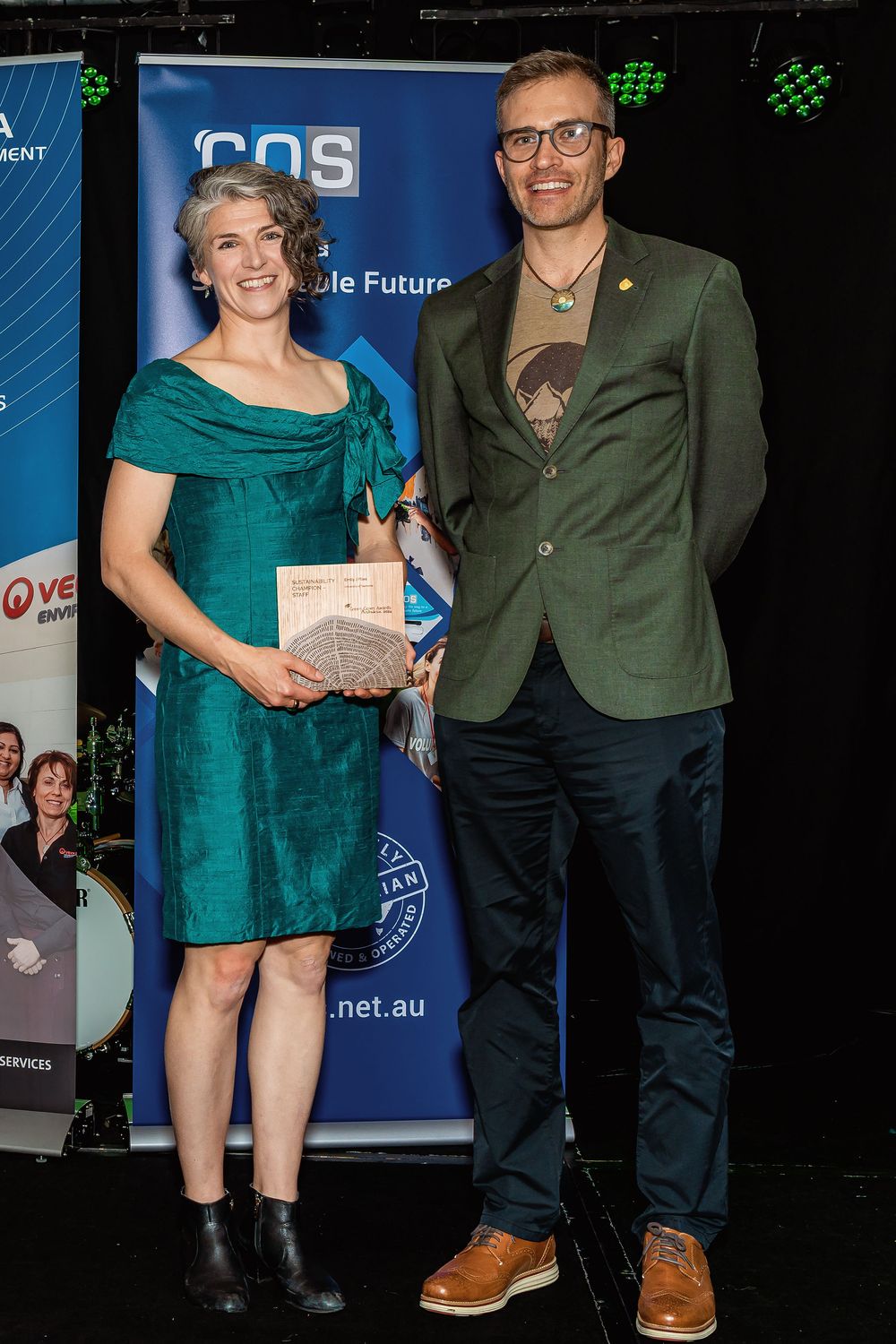 Two people pose on stage at an awards ceremony, woman in teal dress holding a plaque beside man in green blazer.