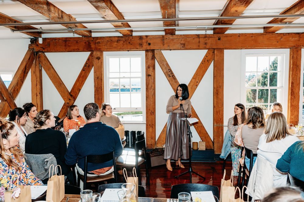 Woman presenting to a seated audience in a rustic wooden beam event space during a workshop or seminar.