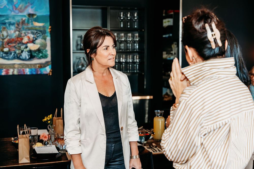 Two women having a conversation at a bar, one in a white blazer and the other in a striped shirt.