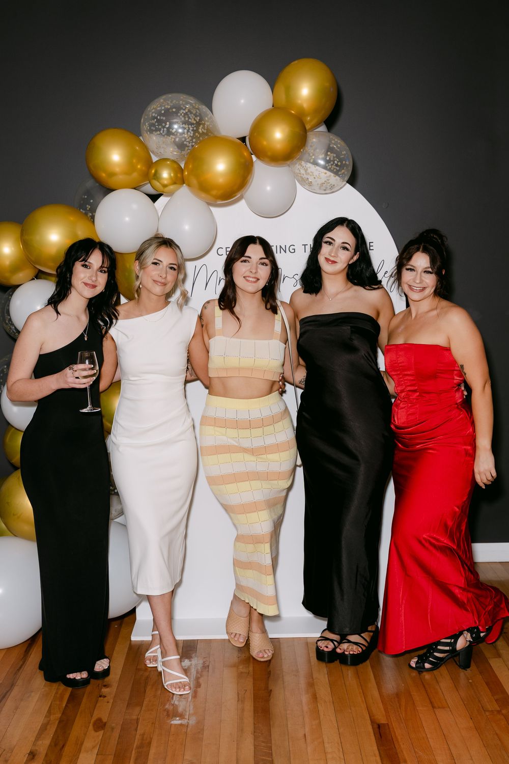 Five women in elegant dresses pose together at a celebration event with gold, white, and silver balloon arch backdrop.